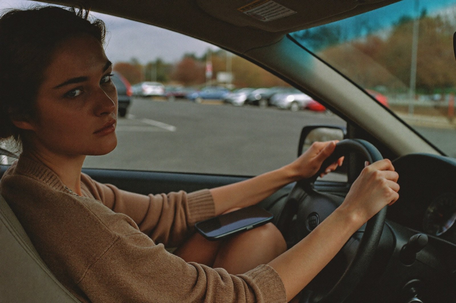 A woman sitting in her parked car in a store parking lot, phone face-down on her lap