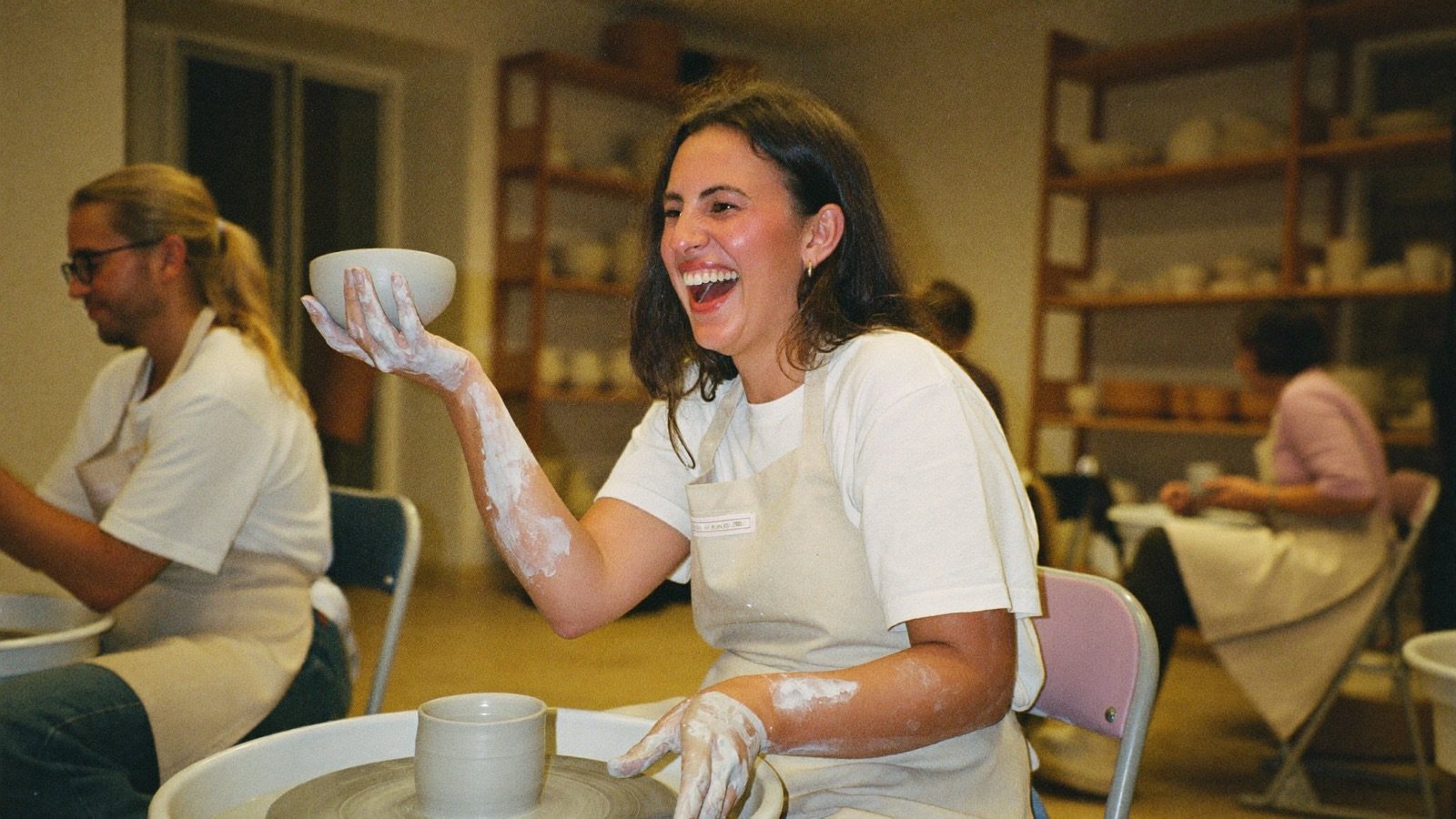 A woman at an evening pottery class, laughing as she holds up the bowl she made